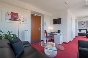 Una habitación de hotel moderna con sofá, una mesa de centro de vidrio, un escritorio con silla, un televisor de pantalla plana y una cama con cabecera.