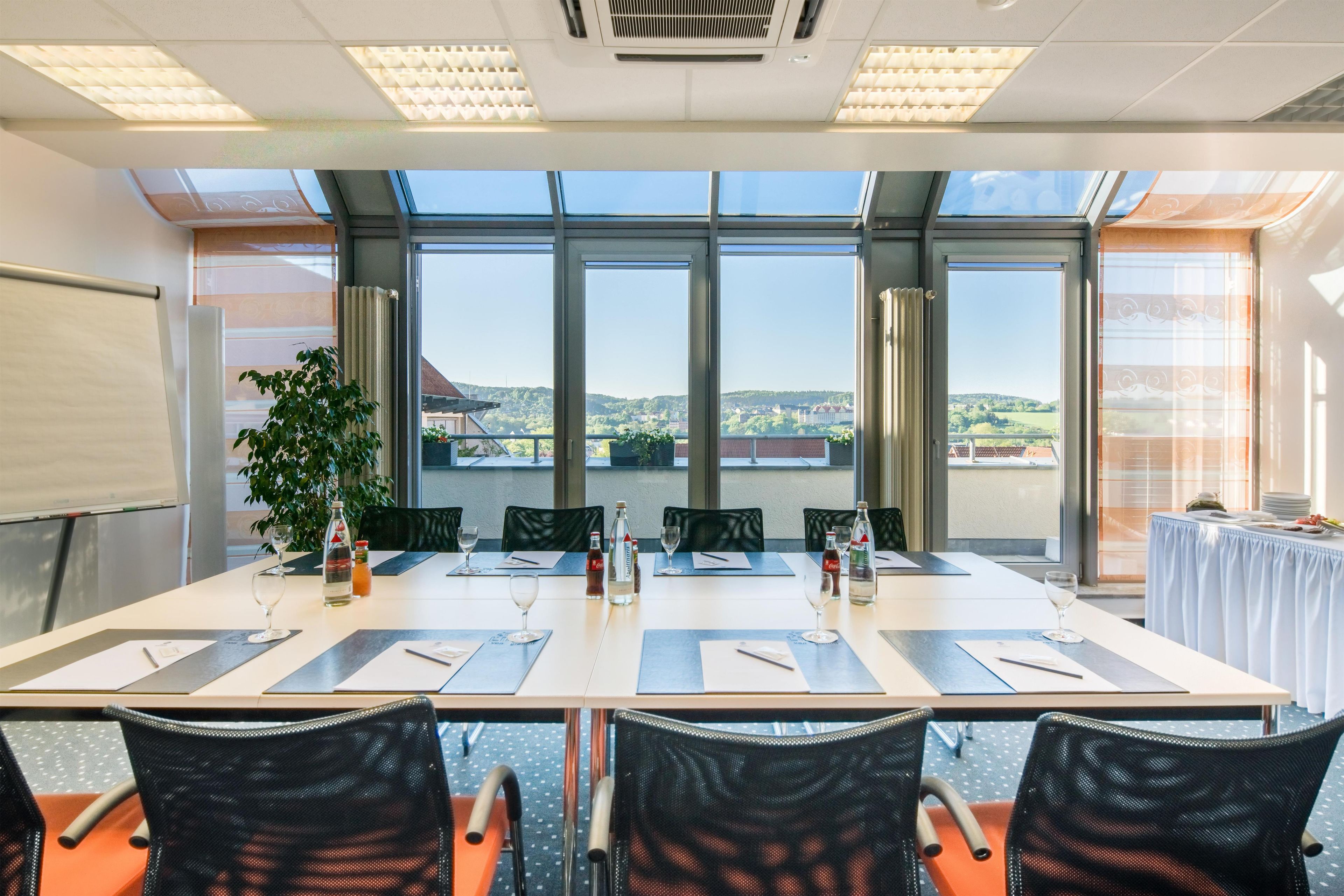 Ein Konferenzraum mit langem Tisch, Stühlen, einer Tafel und Blick auf eine Landschaft durch große Fenster.