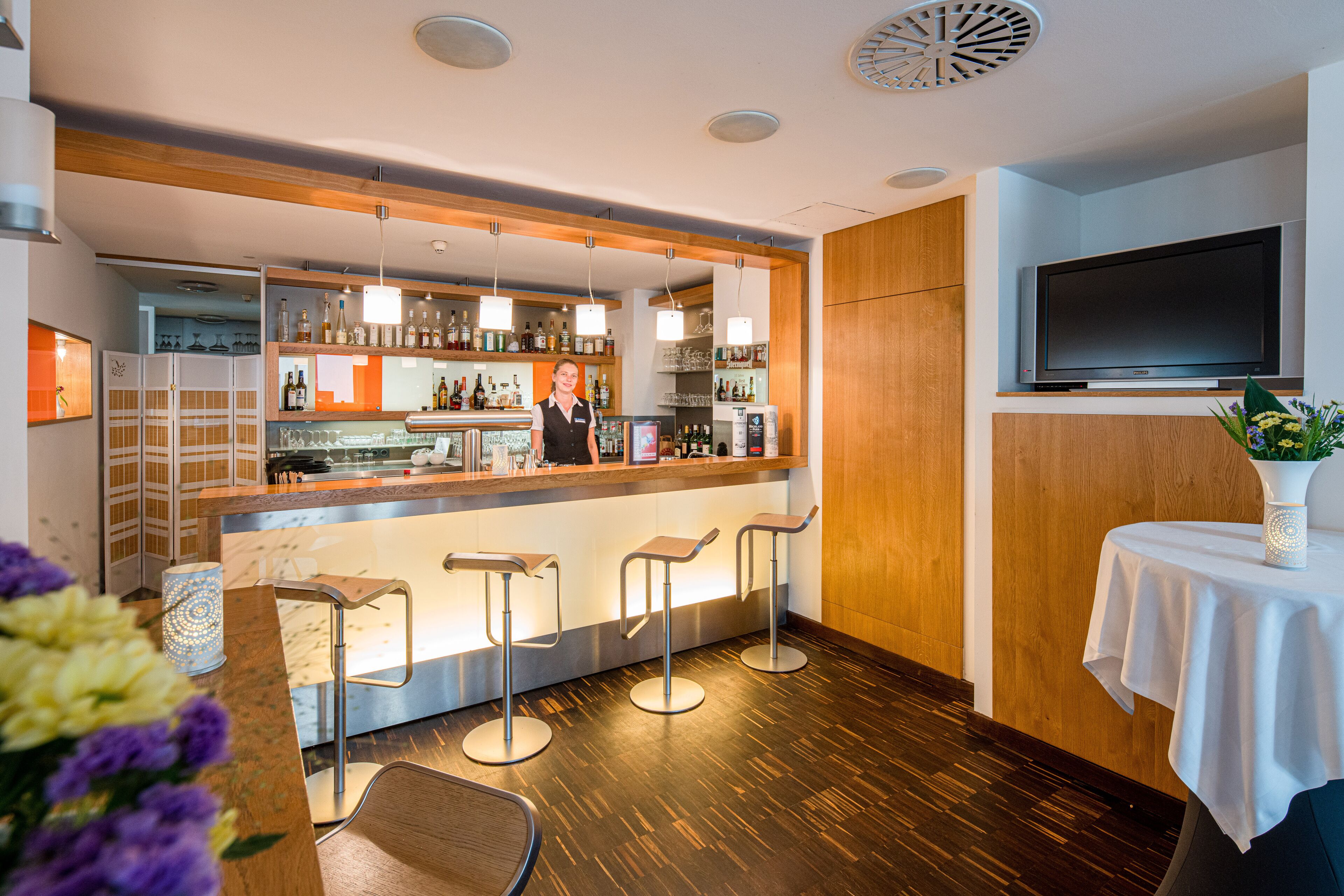 A bar area with a bartender, bar stools, shelves with bottles, and a TV mounted on the wall.