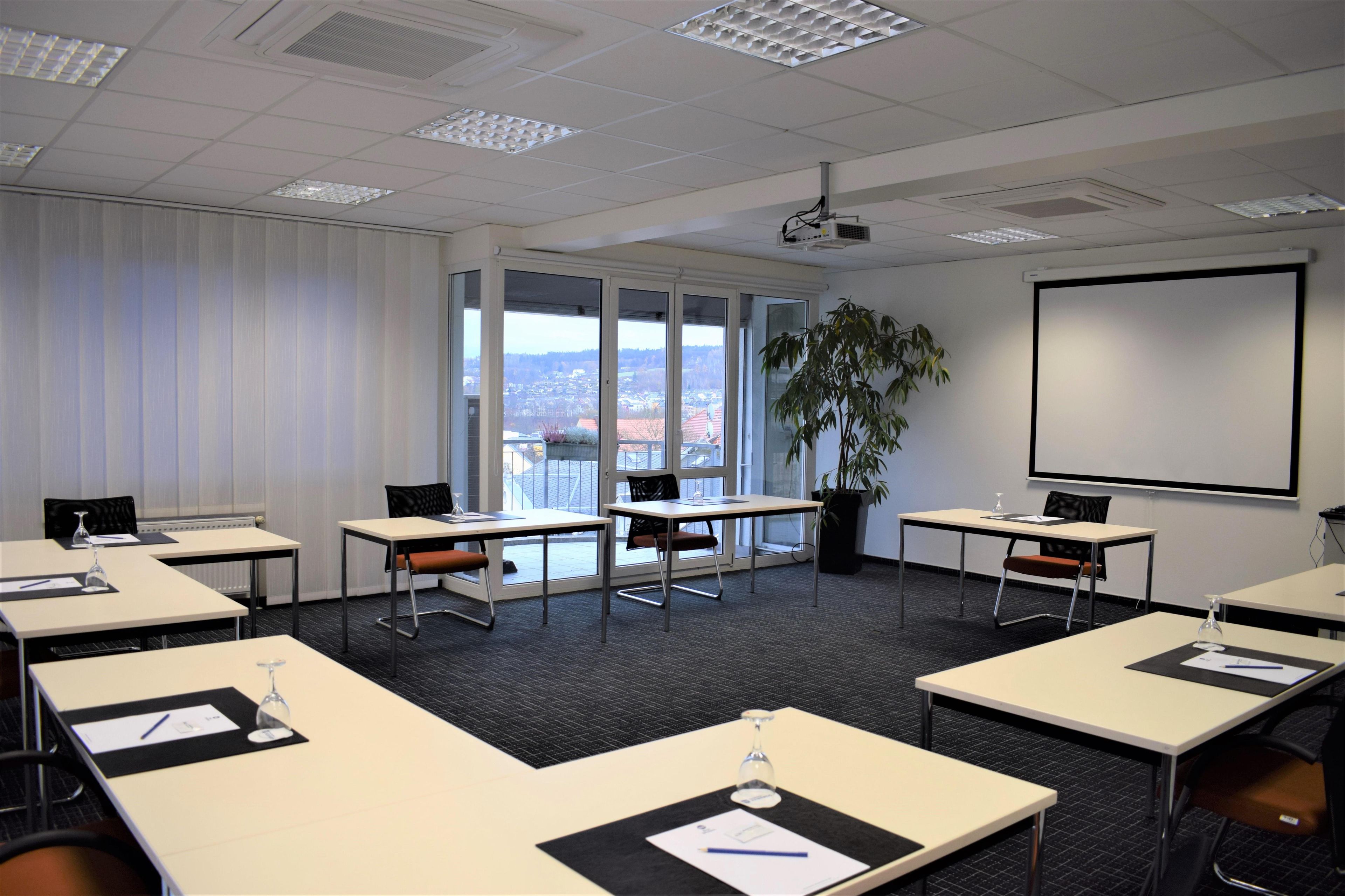 Un salón de conferencias con mesas, sillas, una pantalla de proyector y un amplio ventanal con vista a la ciudad.