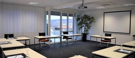 Un salón de conferencias con mesas, sillas, una pantalla de proyector y un amplio ventanal con vista a la ciudad.