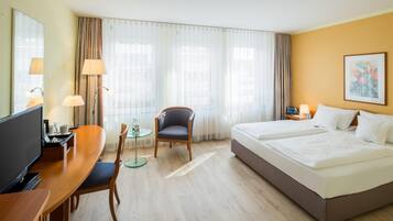 A hotel room with a bed, a desk with a computer, a chair, a TV, and a window with curtains.