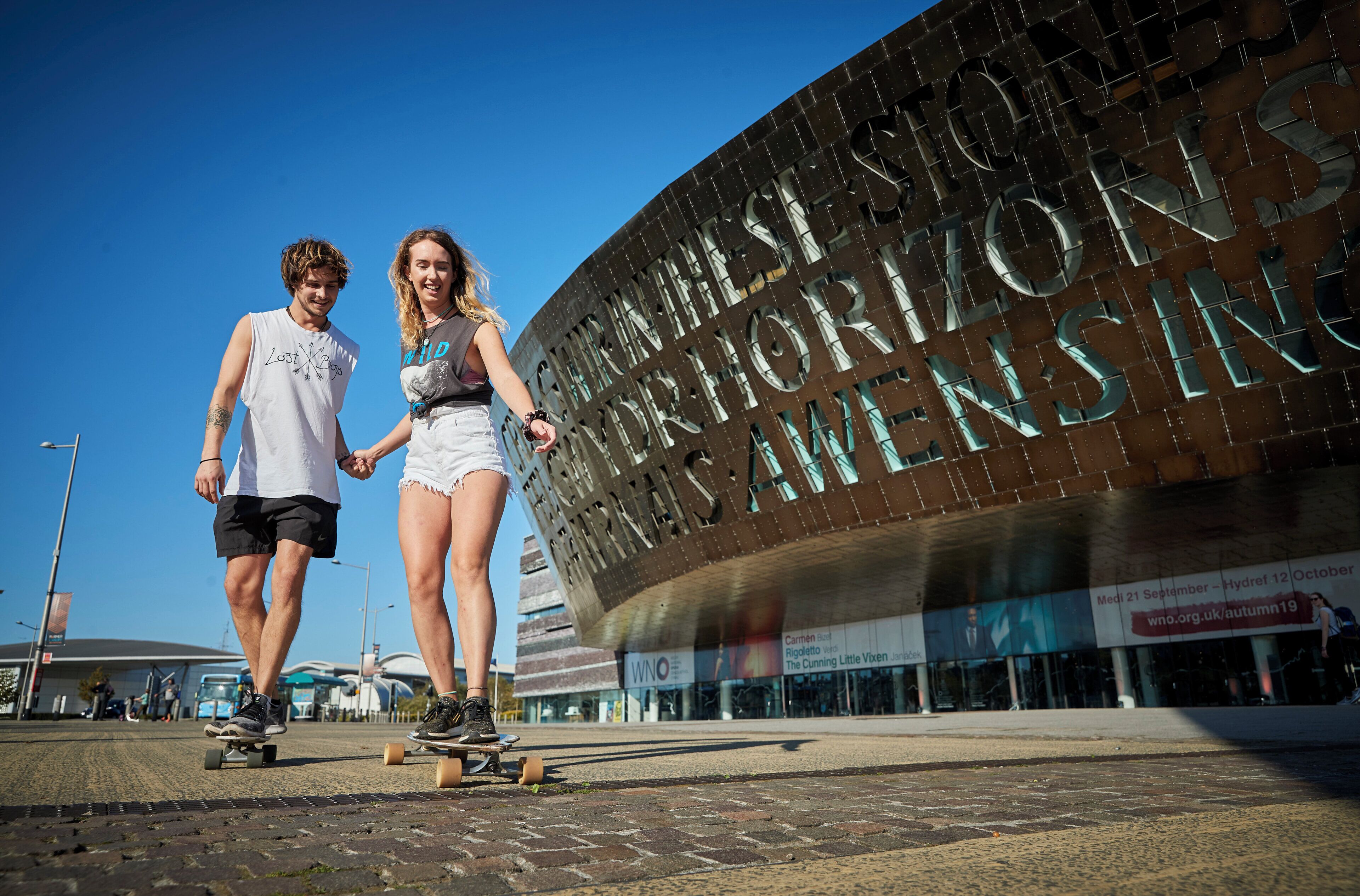 A couple skateboarding in front of a building with large metallic letters.
