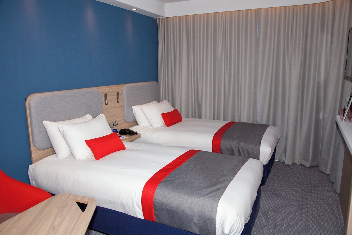 A hotel room with two beds, a wooden headboard, a desk, and a curtain.