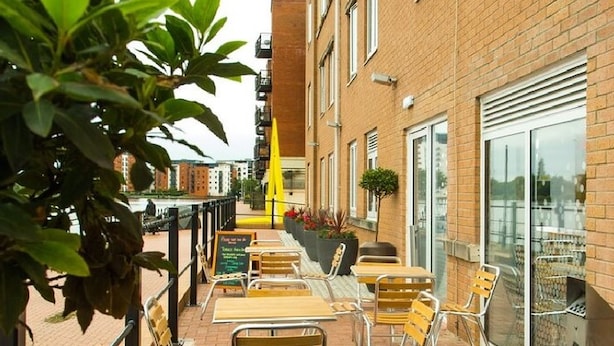 An outdoor seating area with tables and chairs, a yellow umbrella, and a brick building in the background. - Holiday Inn Express Cardiff Bay by IHG (Cardiff)