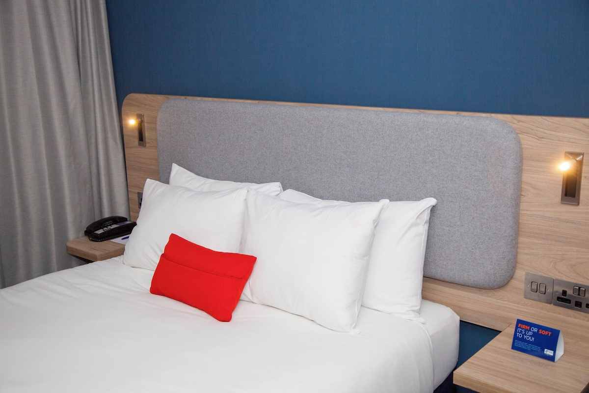A hotel room with a bed, a nightstand with a phone and a book, and a wall-mounted light.