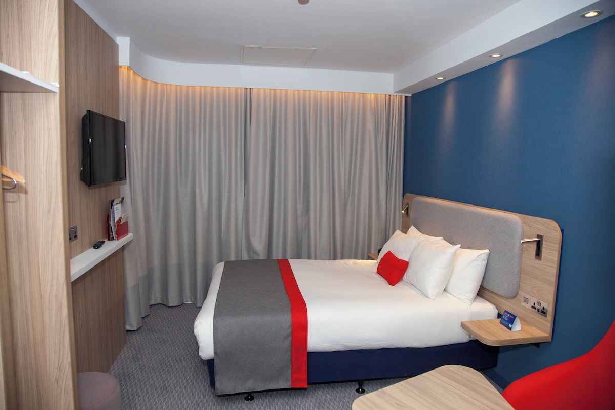 A hotel room with a bed, a TV, a bedside table, and a red chair.