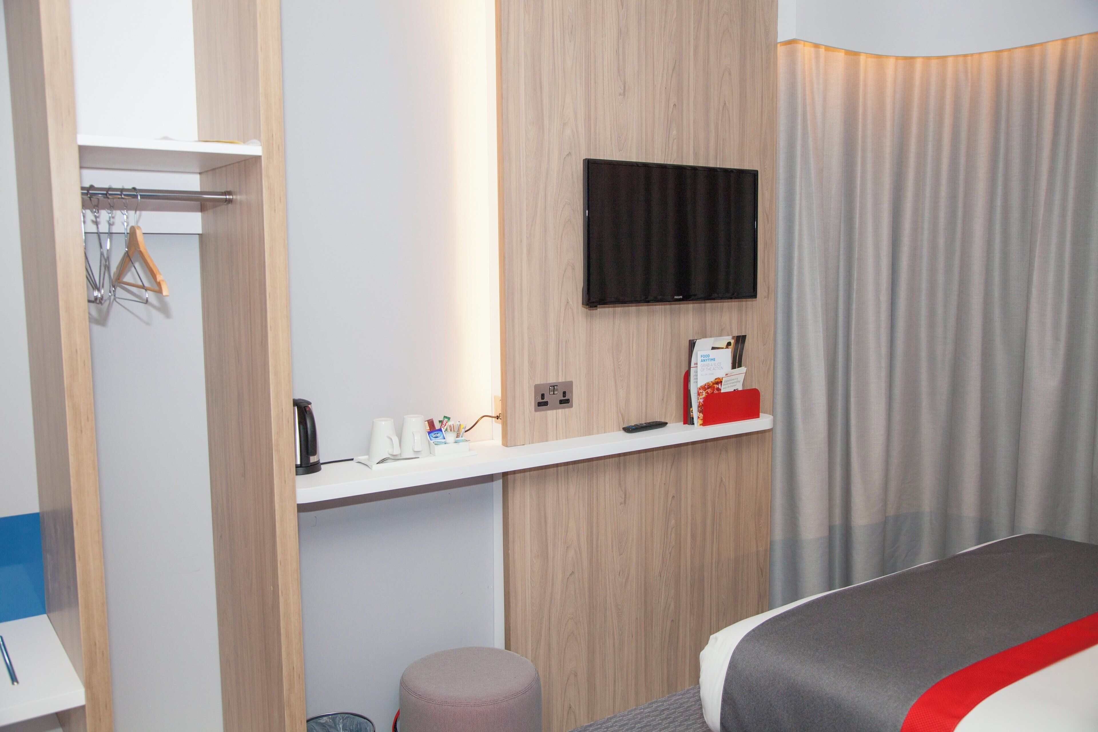 Une chambre d’hôtel avec un lit, un téléviseur fixé au mur, une petite table avec une bouilloire et des tasses, et un rideau.