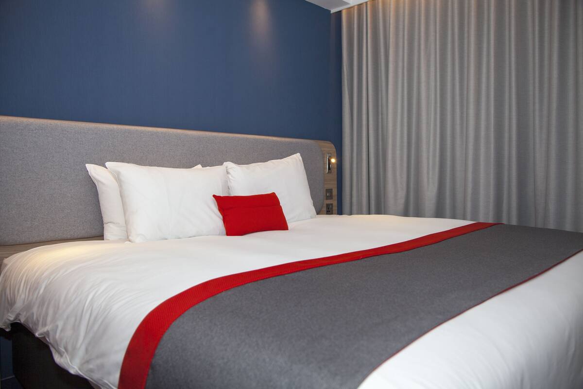 A hotel room with a large bed, grey headboard, white bedding, and a red accent pillow.