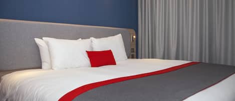 A hotel room with a large bed, grey headboard, white bedding, and a red accent pillow.