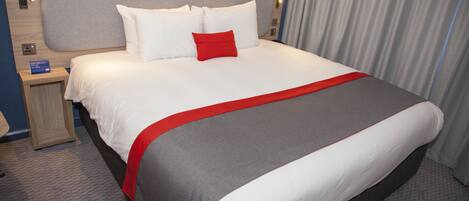 A hotel room with a large bed, a nightstand, a red accent pillow, and a grey and white bedspread.