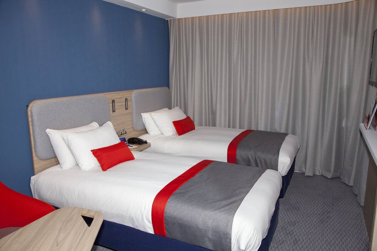 A hotel room with two beds, each with white bedding and red accents, a wooden headboard, a small table, and a curtain.