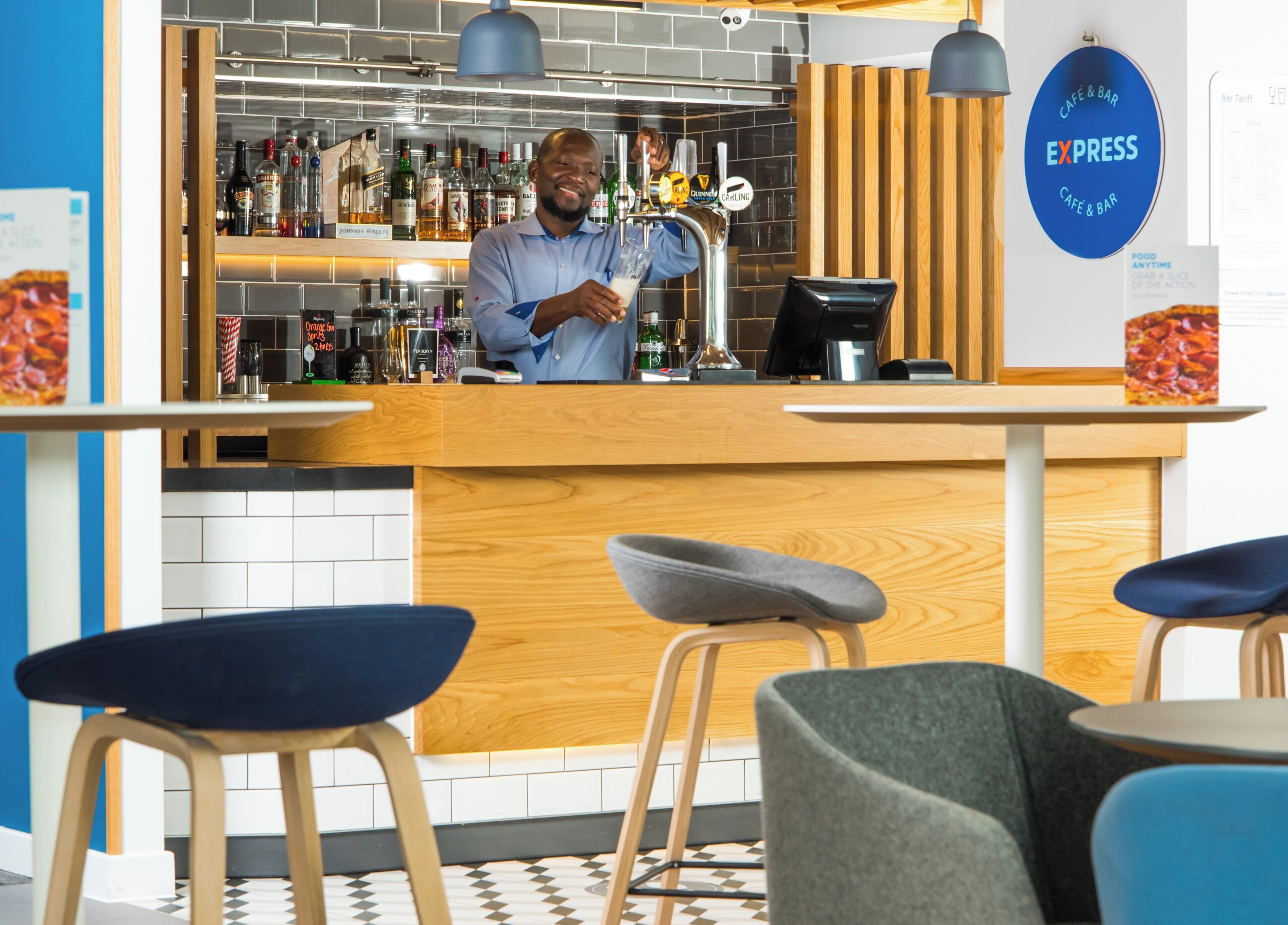 Un espace bar avec un barman, un comptoir où sont exposées différentes bouteilles, et des sièges avec des tabourets.