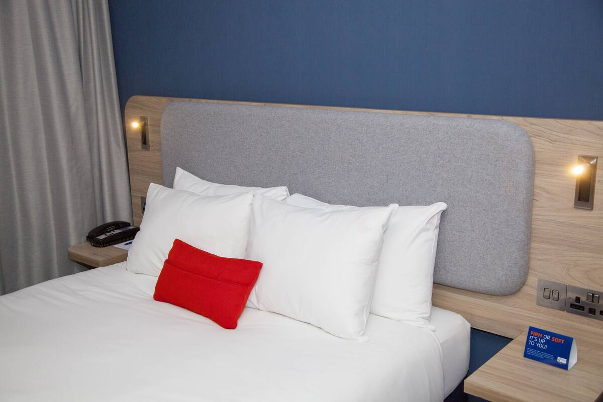 A hotel room with a bed, white pillows, a red pillow, a wooden headboard, a nightstand with a phone and a book, and a blue sign.