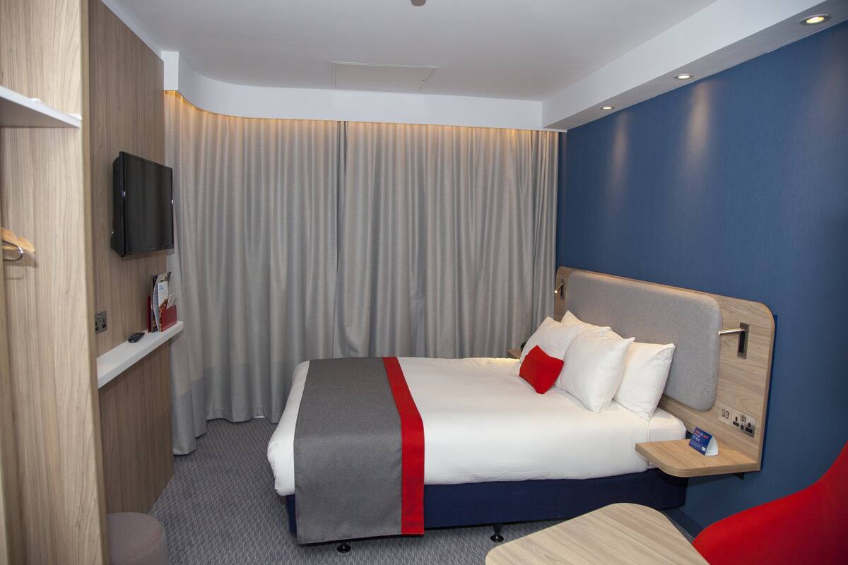 A hotel room with a bed, bedside table, TV, and a red chair.