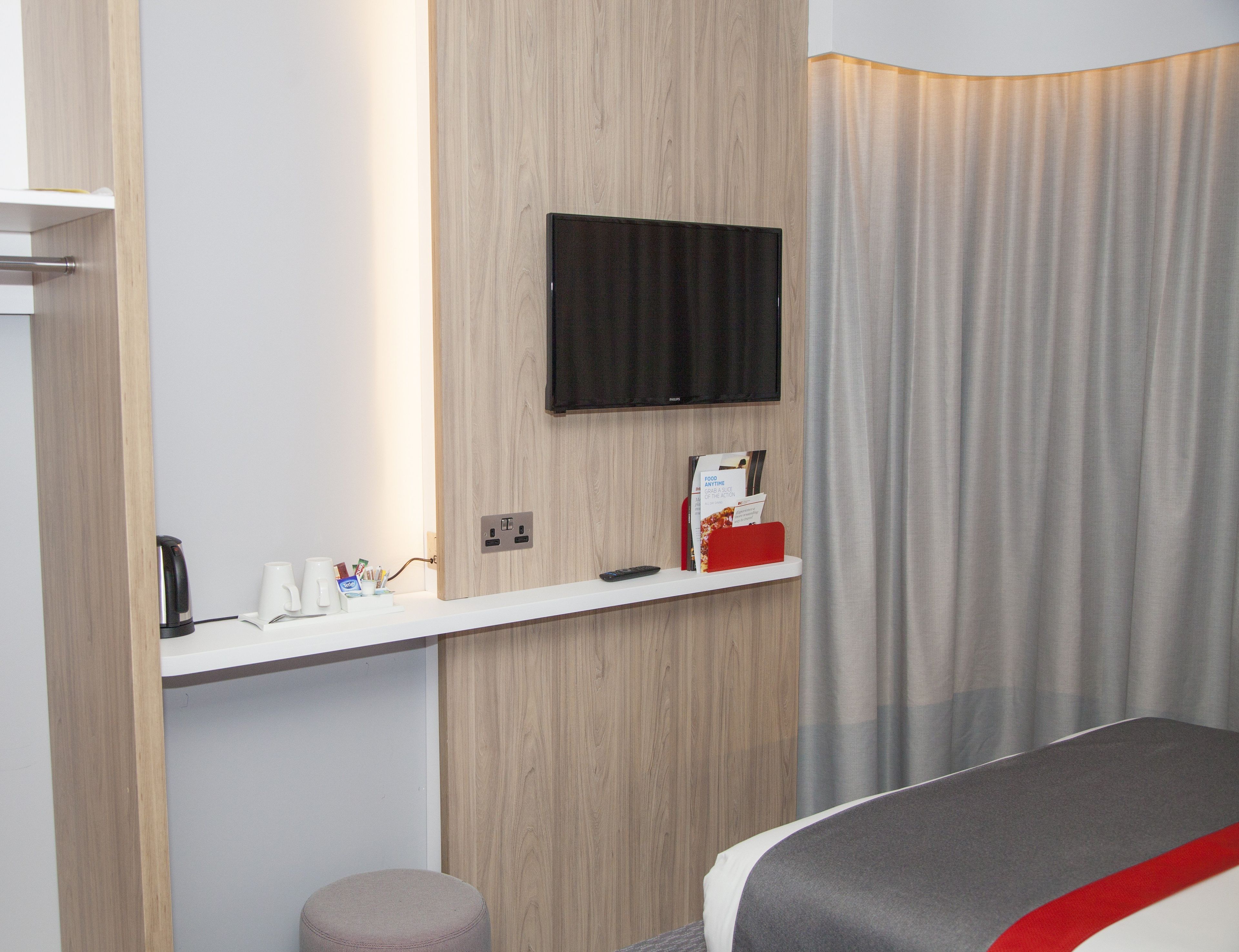 A hotel room with a wooden panel wall, a mounted TV, a bedside table with a red object, and a grey bed.