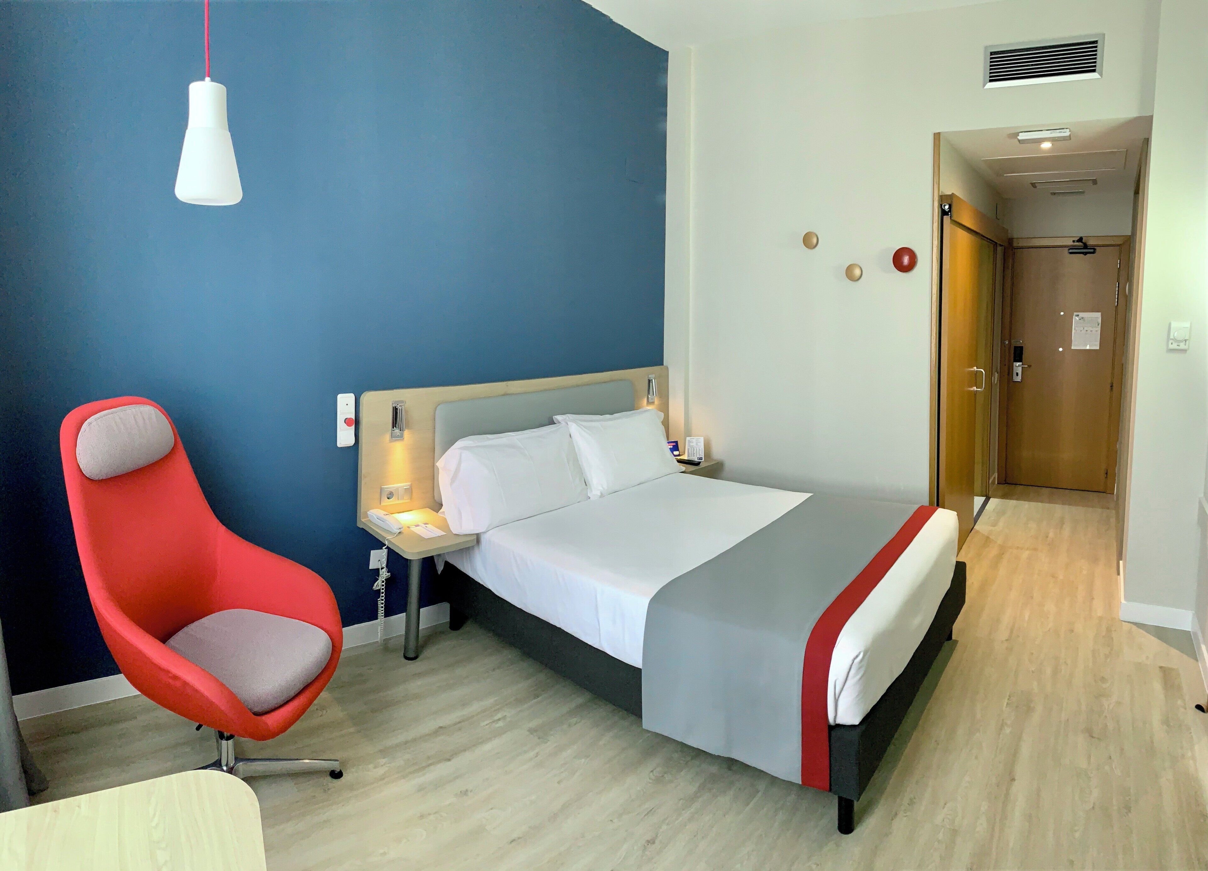 Quarto de hotel com cama, cadeira vermelha, mesa de cabeceira com abajur e porta que dá para outro quarto.
