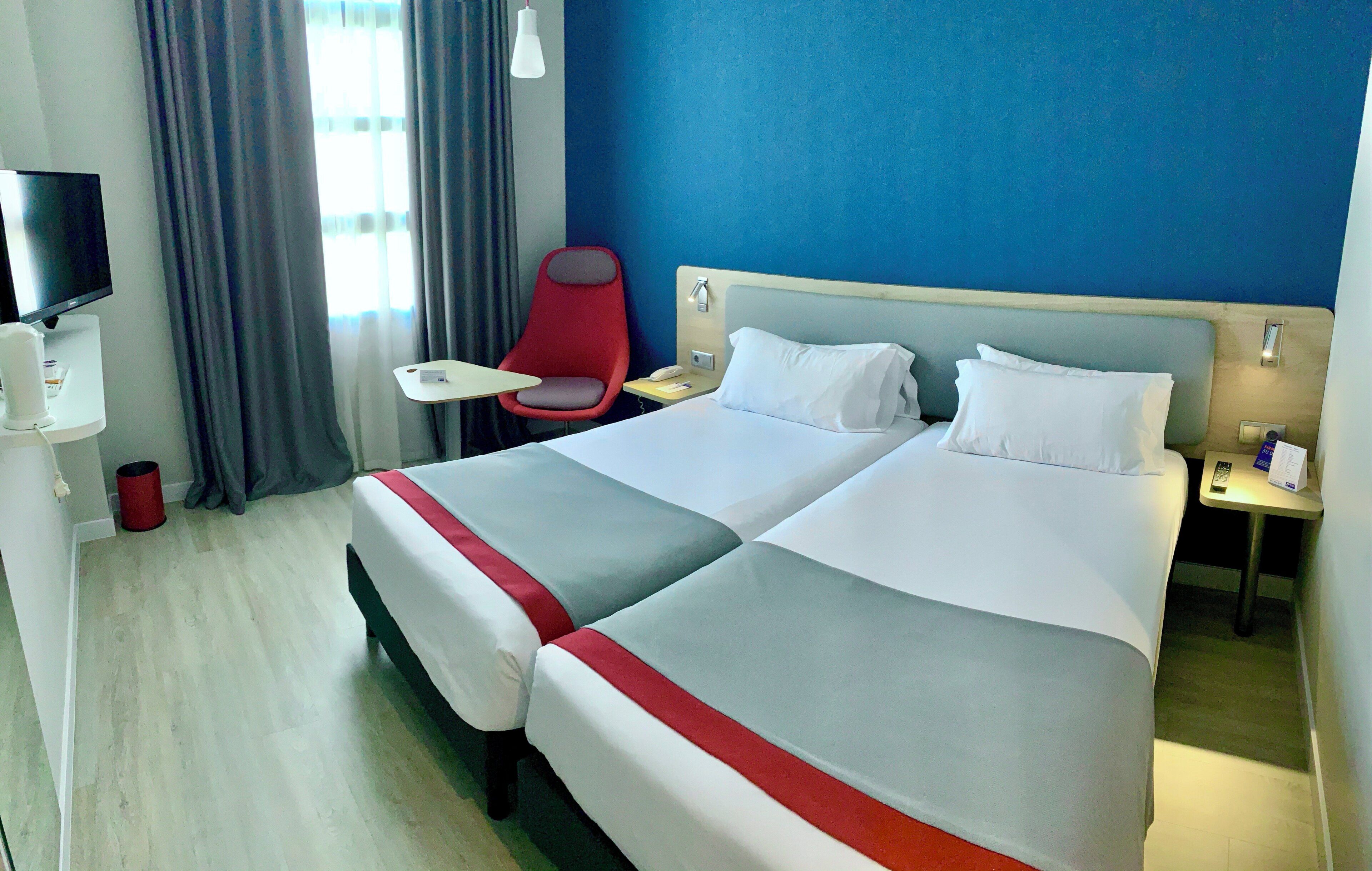 Una habitación de hotel con dos camas, un escritorio, una silla, un televisor y una ventana con cortinas.