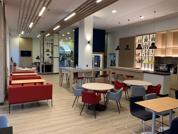 A modern cafe with multiple seating areas, including red and grey chairs, wooden tables, and a bar area with shelves of bottles. - Holiday Inn Express Valencia Ciudad de las Ciencias by IHG (Valencia)