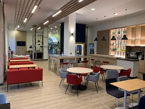 A modern cafe with multiple seating areas, including red and grey chairs, wooden tables, and a bar area with shelves of bottles. - Holiday Inn Express Valencia Ciudad de las Ciencias by IHG (Valencia)