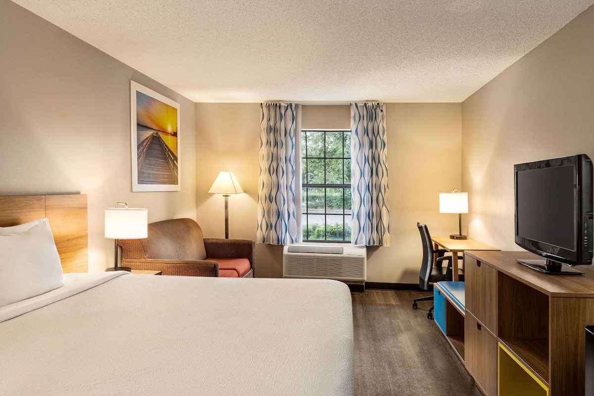 A hotel room with a bed, two chairs, a desk, a television, and a window with curtains.