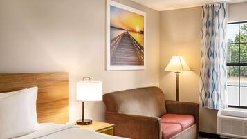 A hotel room with a bed, a chair, a nightstand with a lamp, a painting of a sunset over a pier, and a window with curtains.