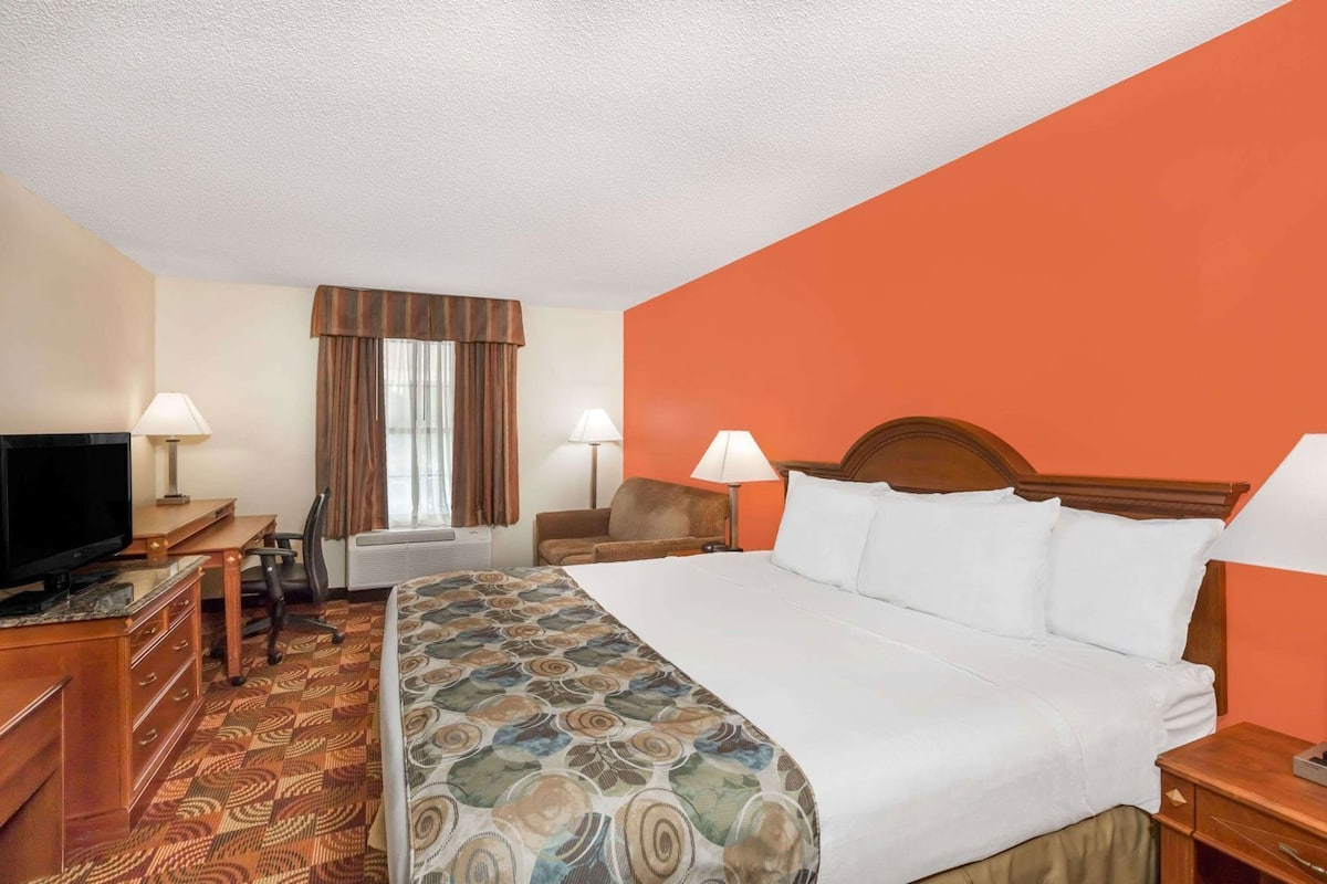 A hotel room with a bed, a desk with a TV, two chairs, a window with curtains, and a lamp.