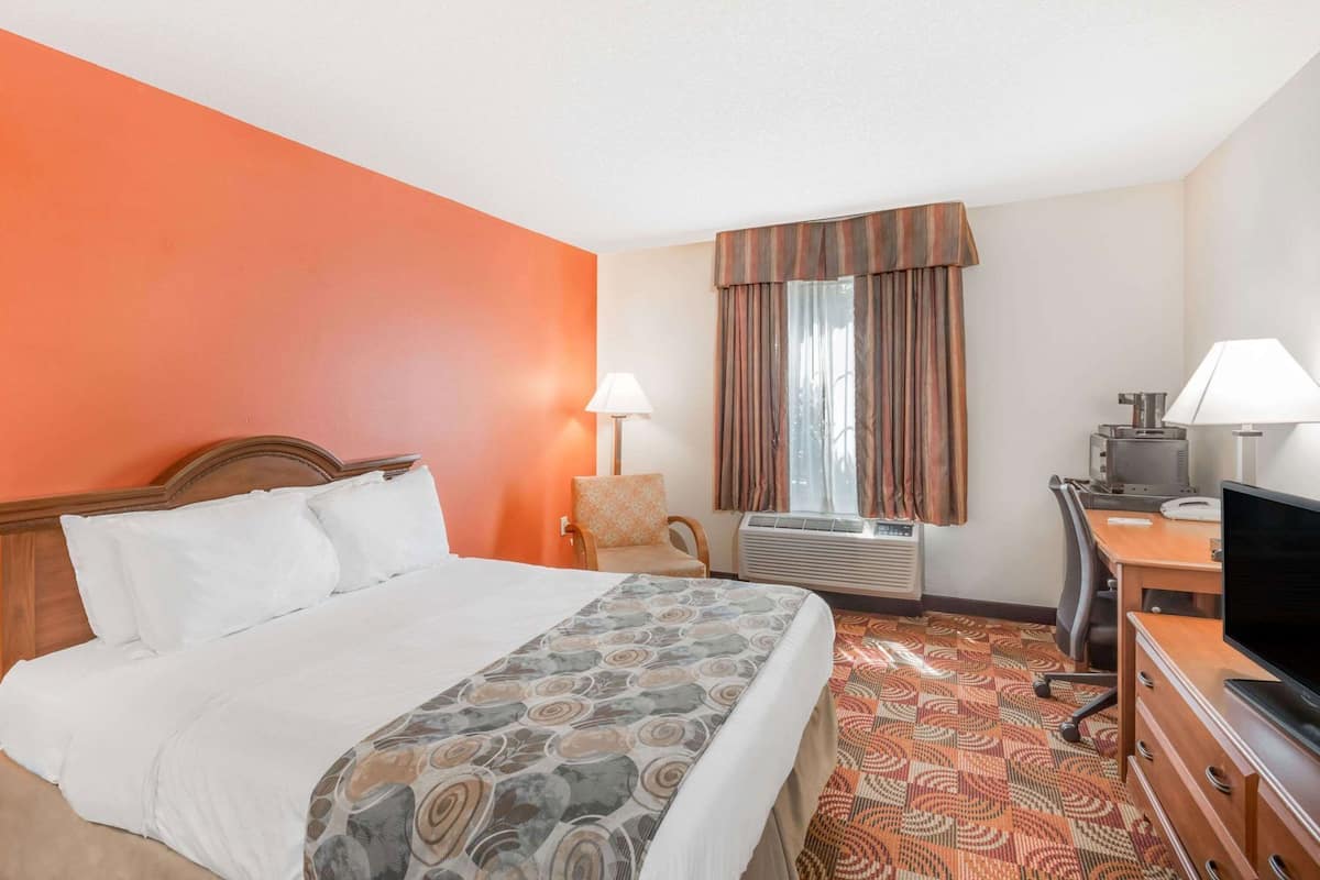 A hotel room with a bed, desk, chair, TV, lamp, air conditioning unit, and a window with curtains.