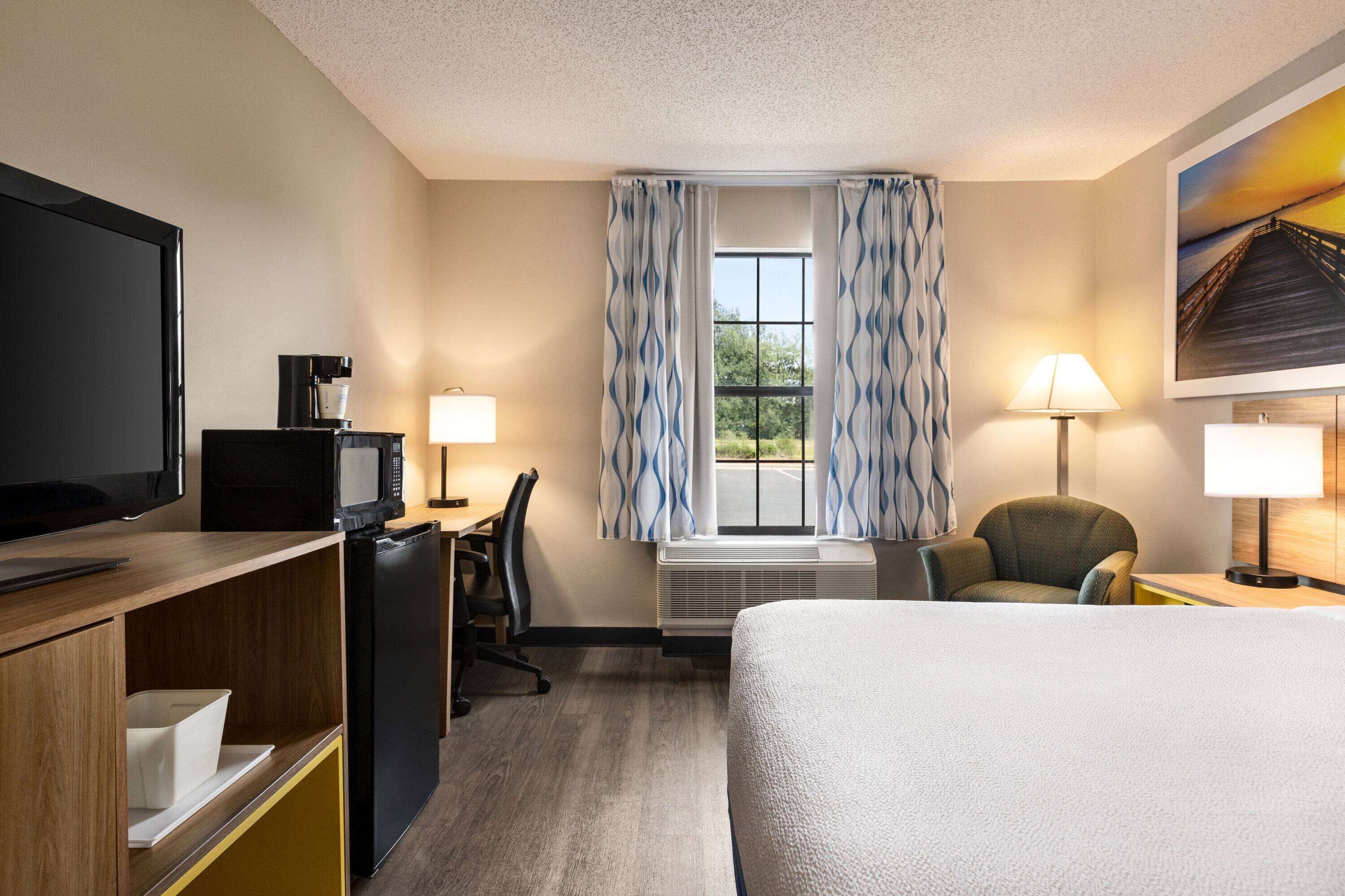 A hotel room with a bed, a desk with a computer, a television, a coffee maker, and a window with curtains.