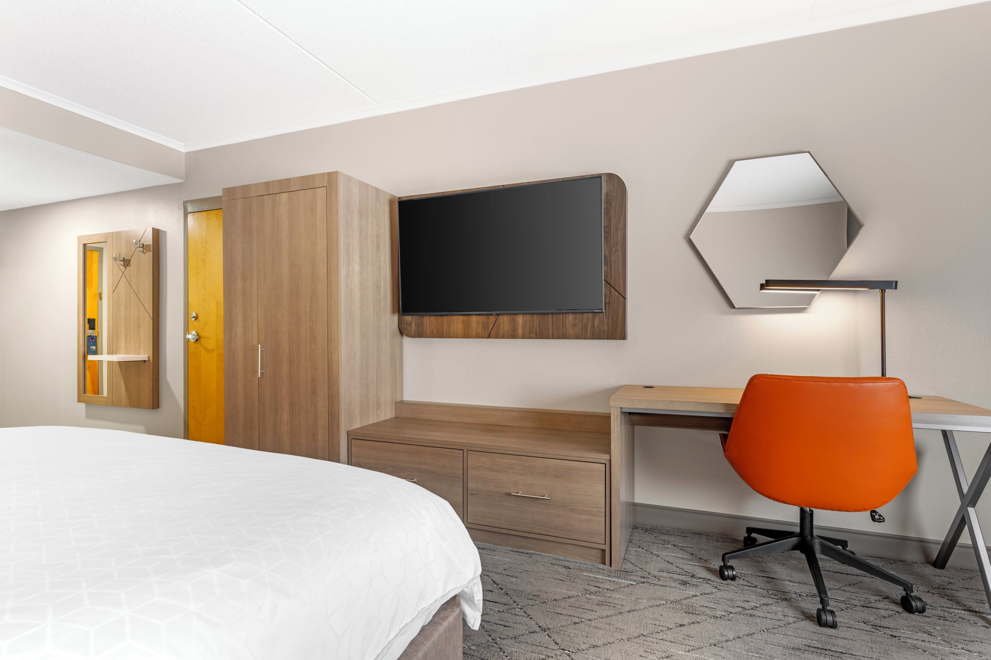 A hotel room with a bed, a desk, an orange chair, a wardrobe, and a flat-screen TV.