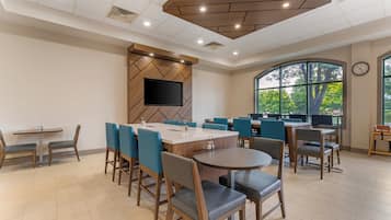 A modern dining area with multiple tables and chairs, a flat-screen TV mounted on a wooden wall, and large windows providing natural light.