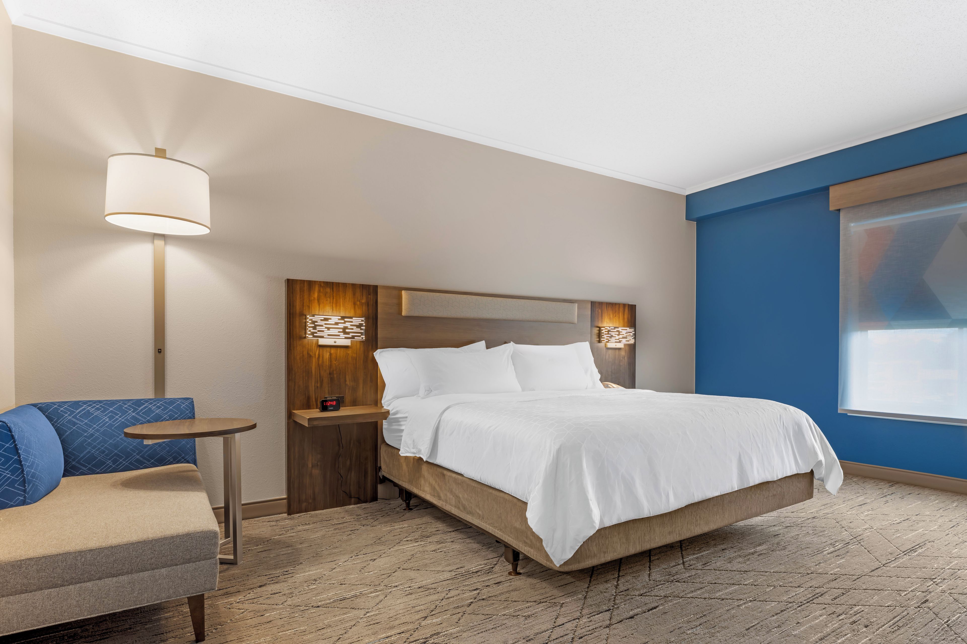 Habitación de hotel con cama, una silla, mesita de noche, lámpara y un reloj de pared.
