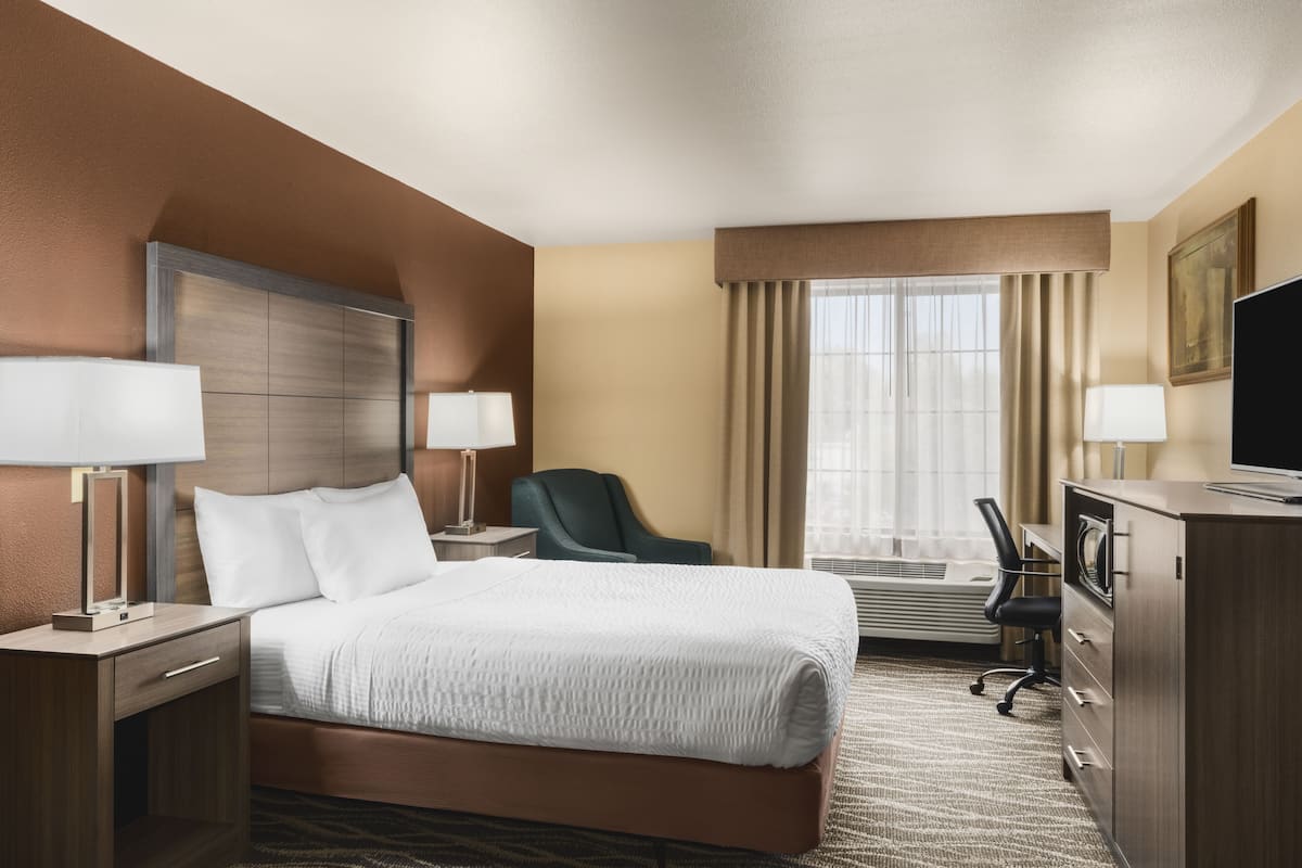 A hotel room with a large bed, two bedside tables with lamps, a desk with a chair, a television, and a window with curtains.
