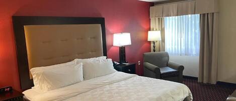 A hotel room with a large bed, a nightstand, a chair, a lamp, and a window with curtains.