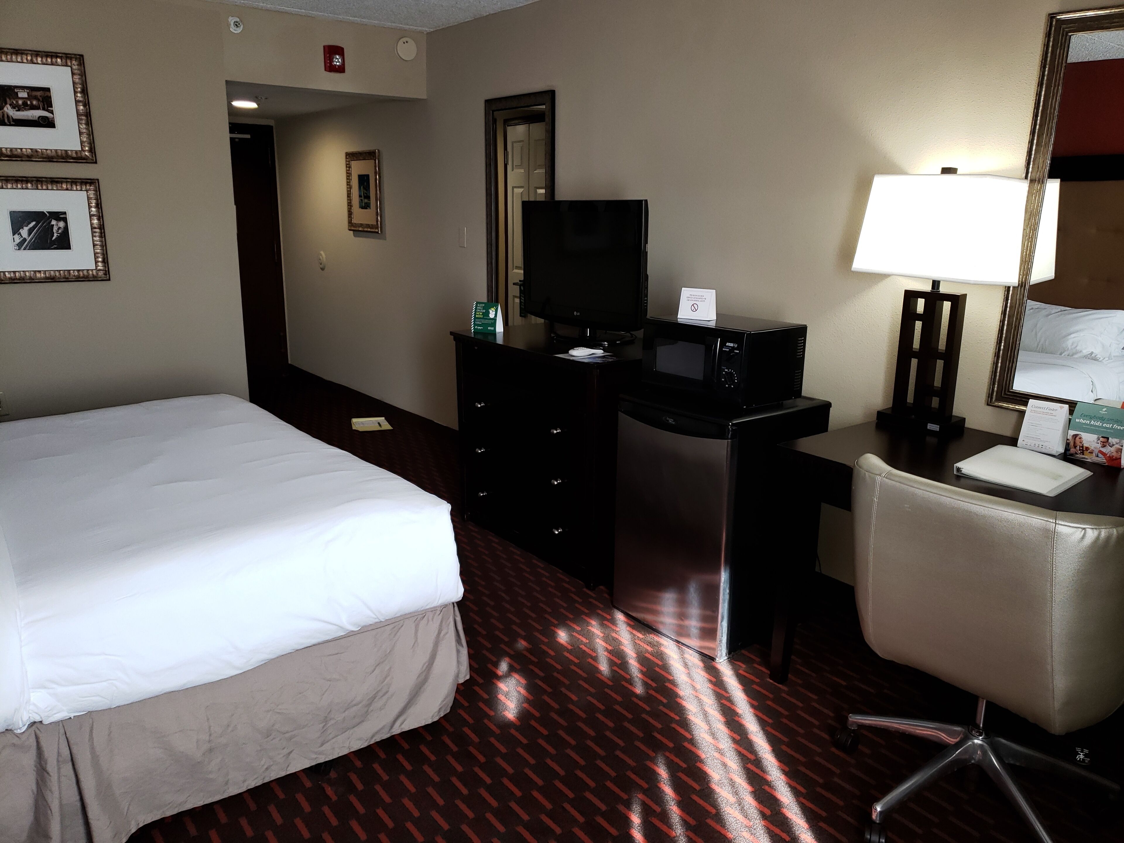 A hotel room with a bed, a desk with a computer and a TV, a chair, and a lamp.