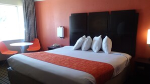 Ein Hotelzimmer mit einem großen Bett, einem Nachttisch mit Telefon, einem kleinen runden Tisch und zwei orangefarbenen Stühlen sowie einem Fenster mit Vorhängen.