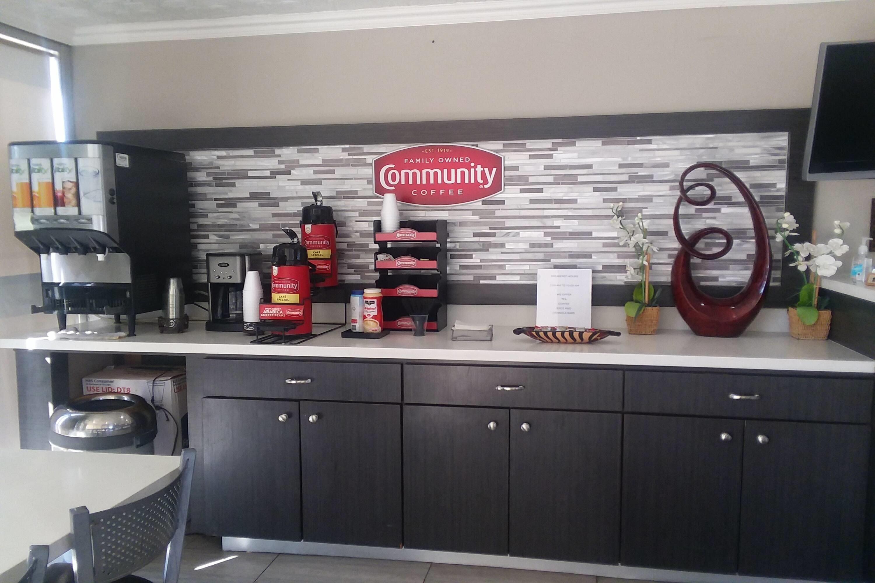 A coffee station with a coffee machine, milk frother, and various coffee pods. There is a sign that reads "Community Coffee" and decorative plants.