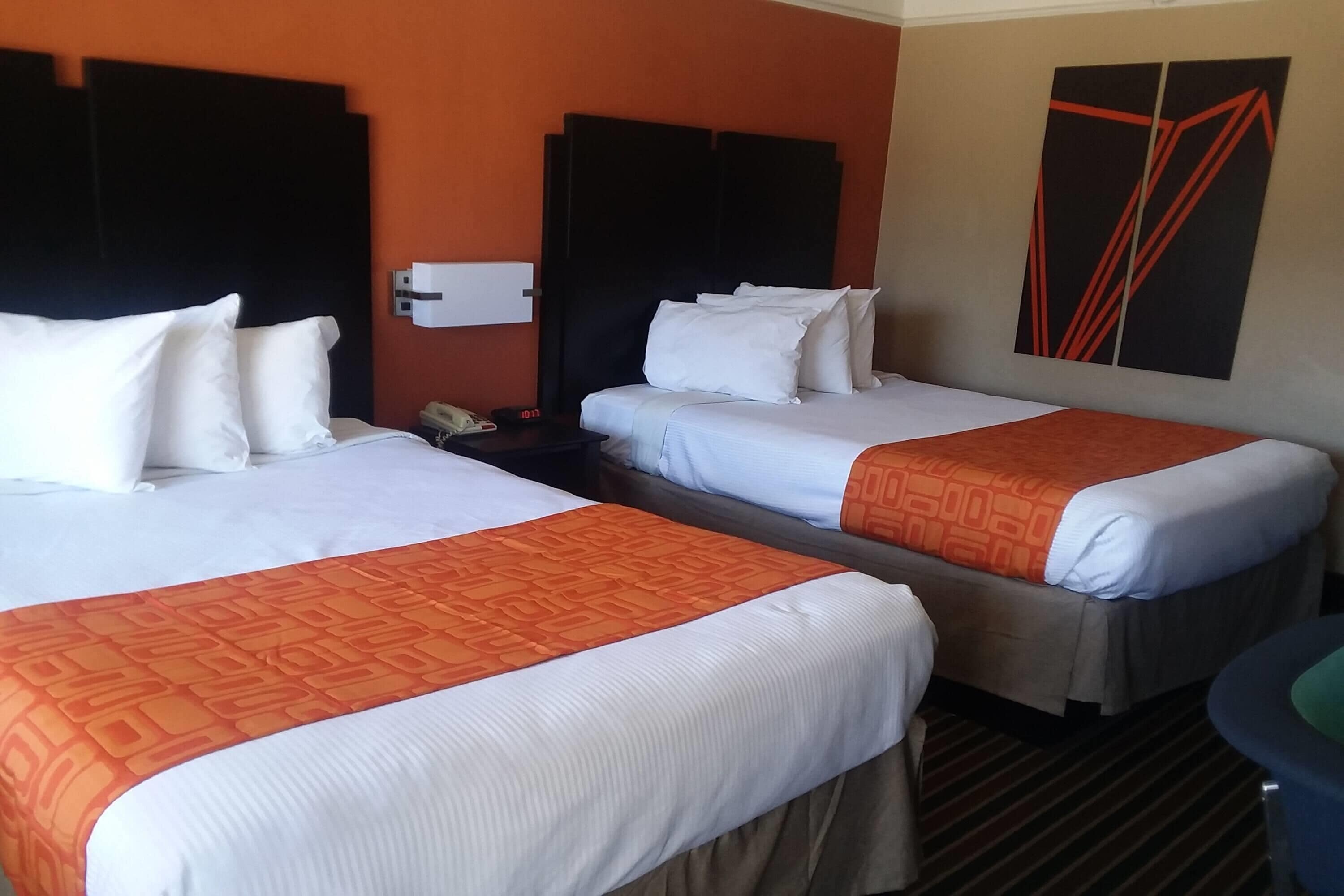 A hotel room with two beds, each with white bedding and orange accents. There is a wall-mounted air conditioning unit and a painting above the beds.