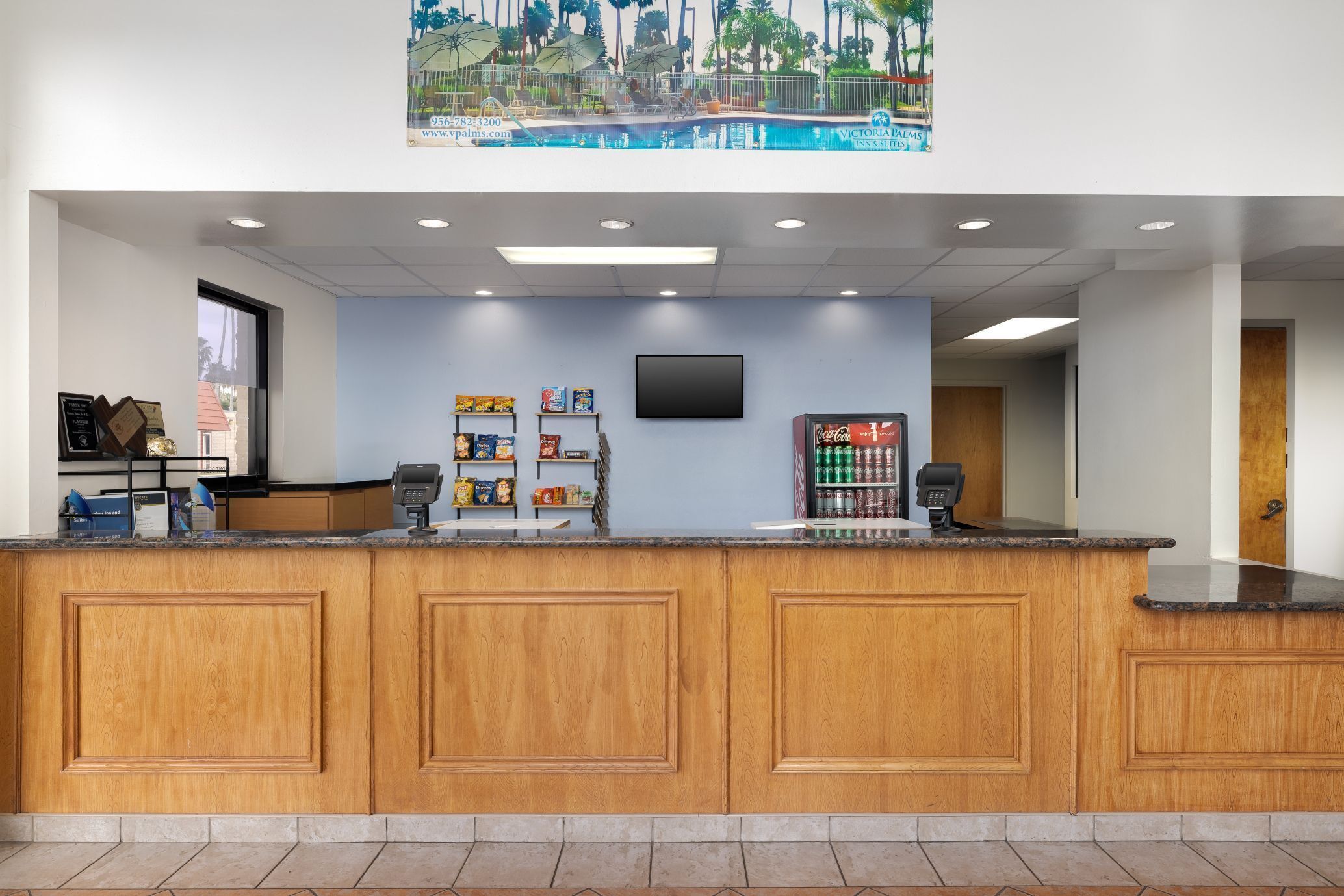 A reception area with a wooden counter, a flat-screen TV mounted on the wall, and a window with a view of palm trees.