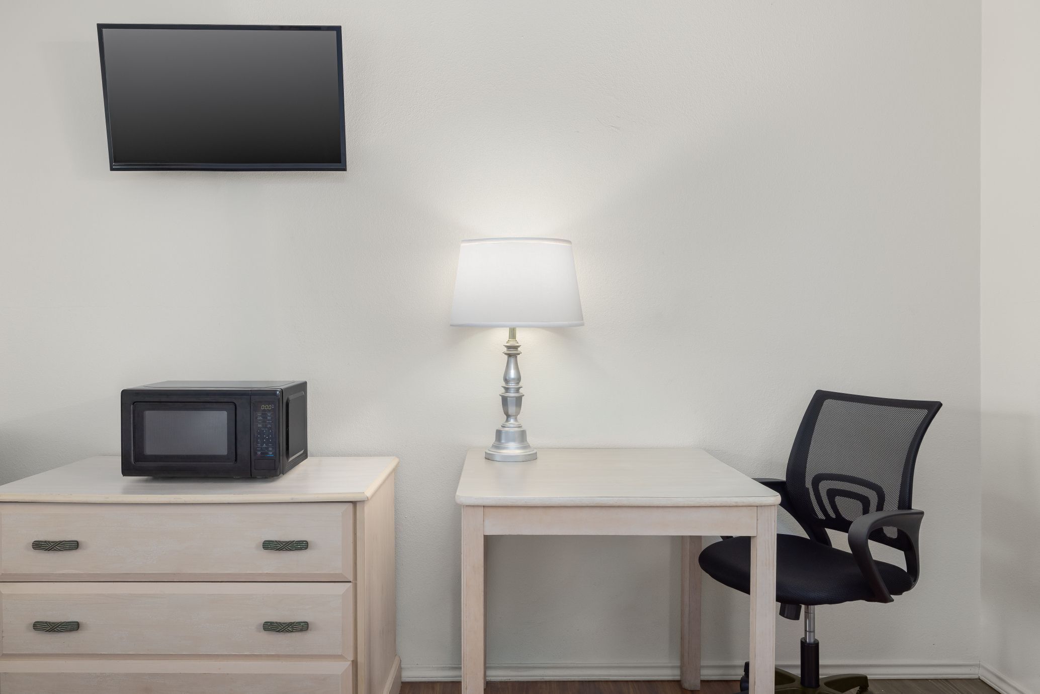 Una habitación pequeña con microondas en un mueble cajón, una mesa con lámpara y una silla.