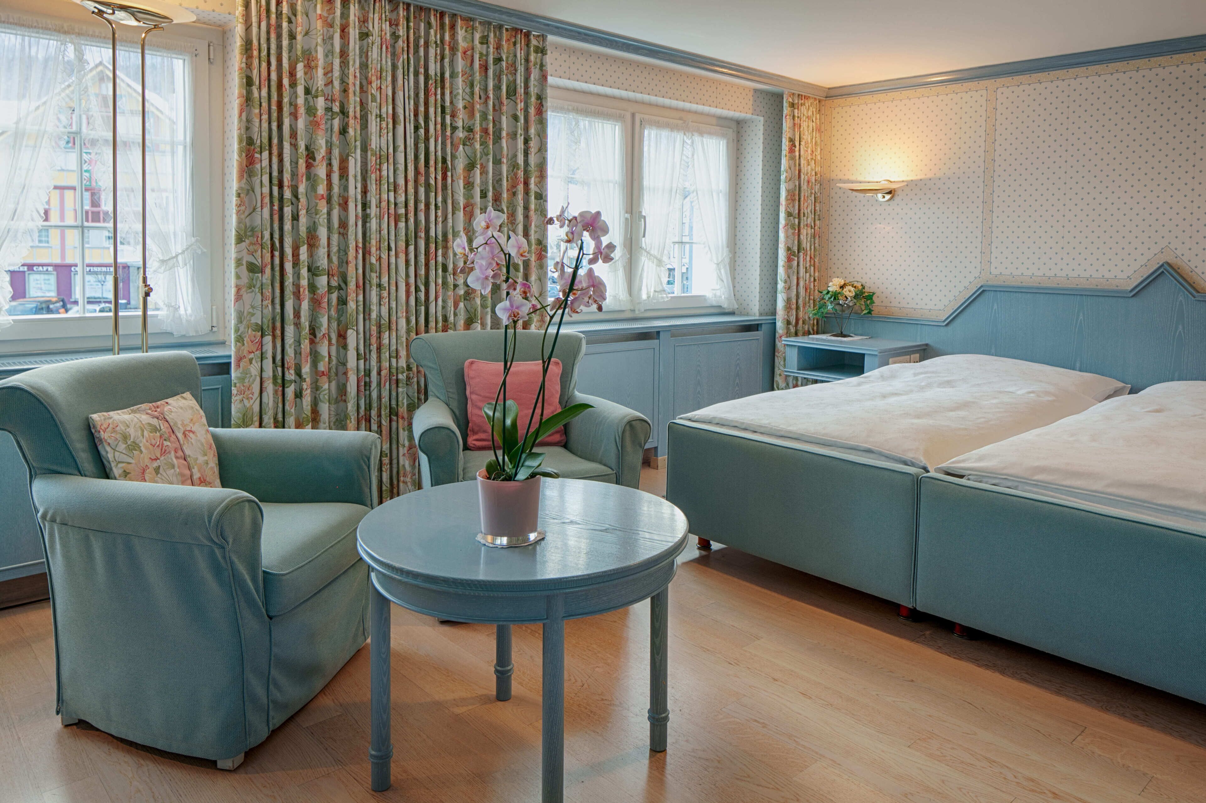 Habitación de hotel con cama, dos sillones, una mesa redonda con planta y una ventana con cortinas.