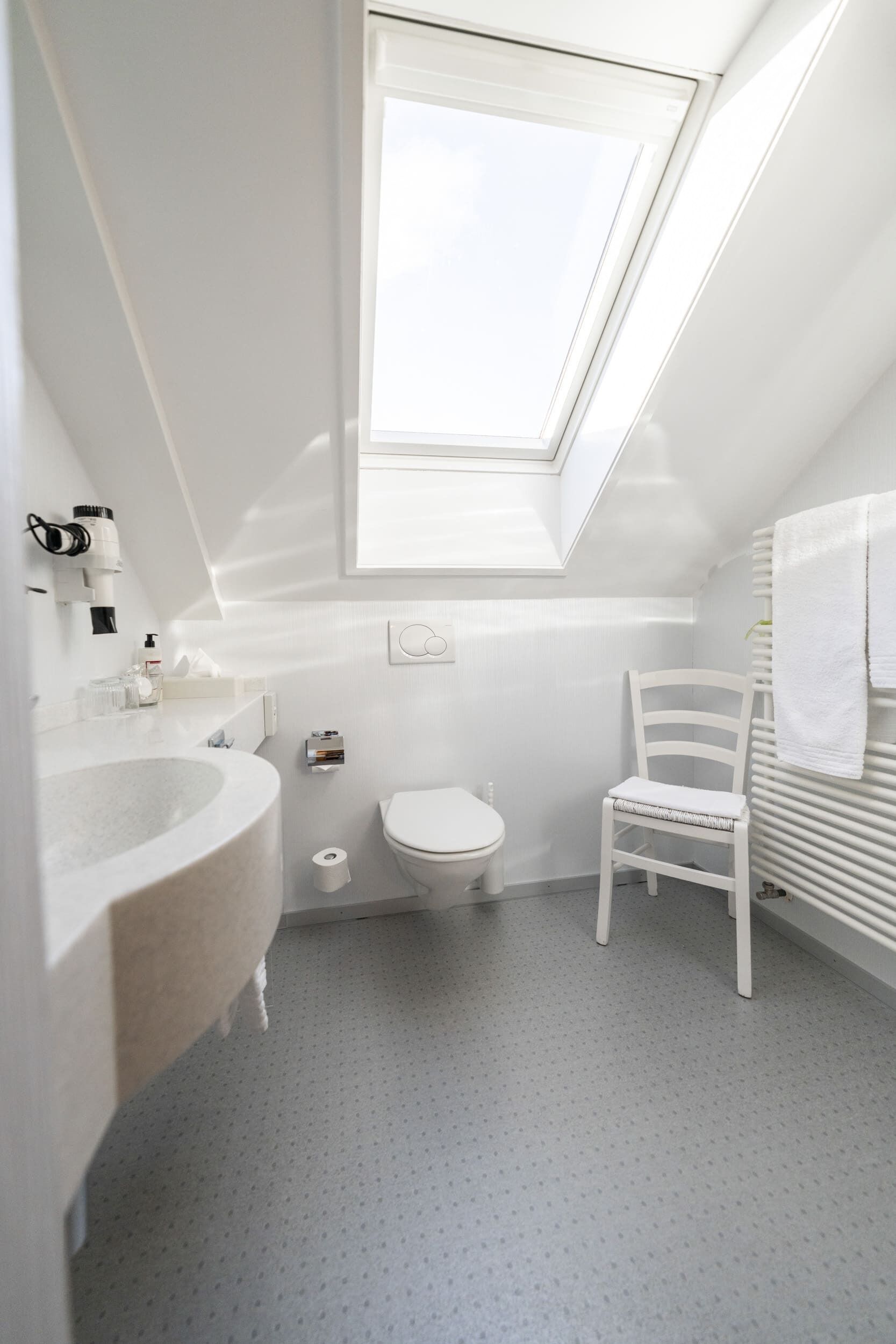 A small, white bathroom with a skylight, a white sink, a toilet, and a white chair.