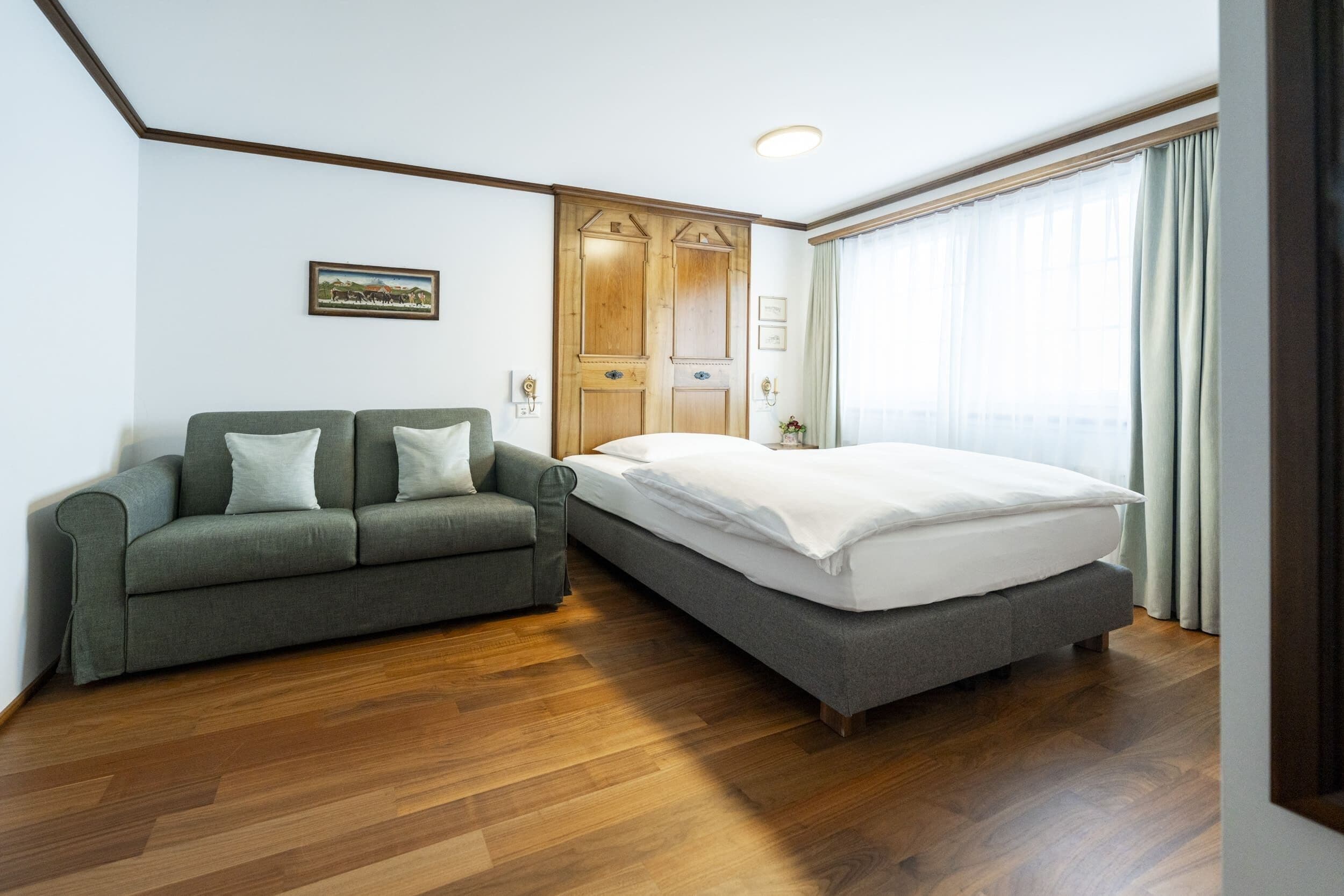 A bedroom with a bed, a sofa, wooden flooring, a wooden wardrobe, and a window with curtains.