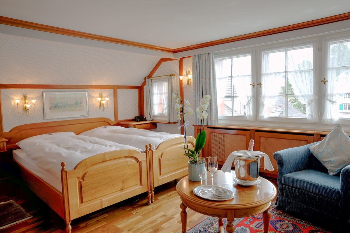 Habitación de hotel con dos camas de madera, una mesa de comedor con una jofaina y vasos, un sillón azul y ventanas grandes con cortinas.