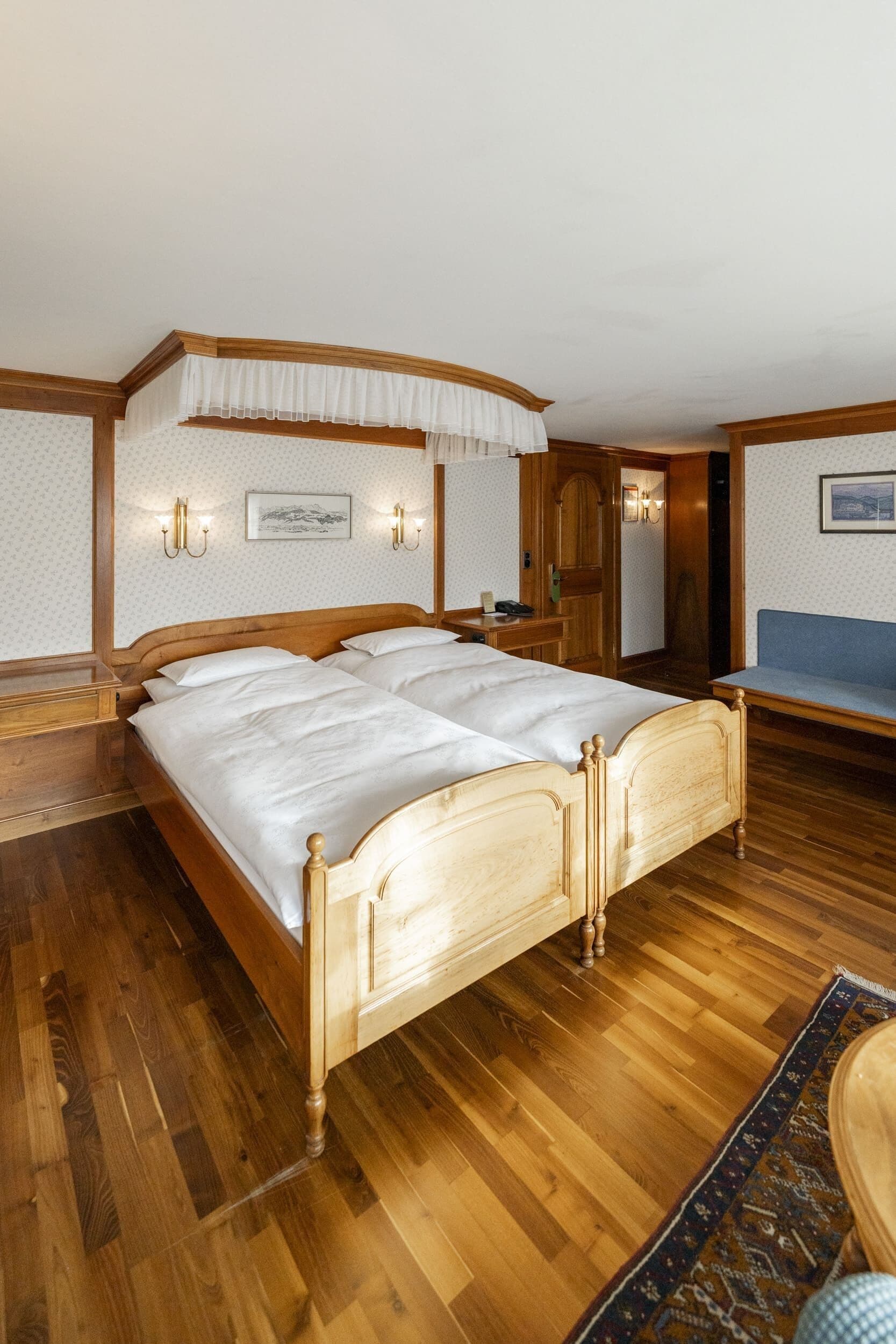 Un lit à baldaquin en bois, agrémenté de draps blancs, un banc en bois et un tapis à motifs.