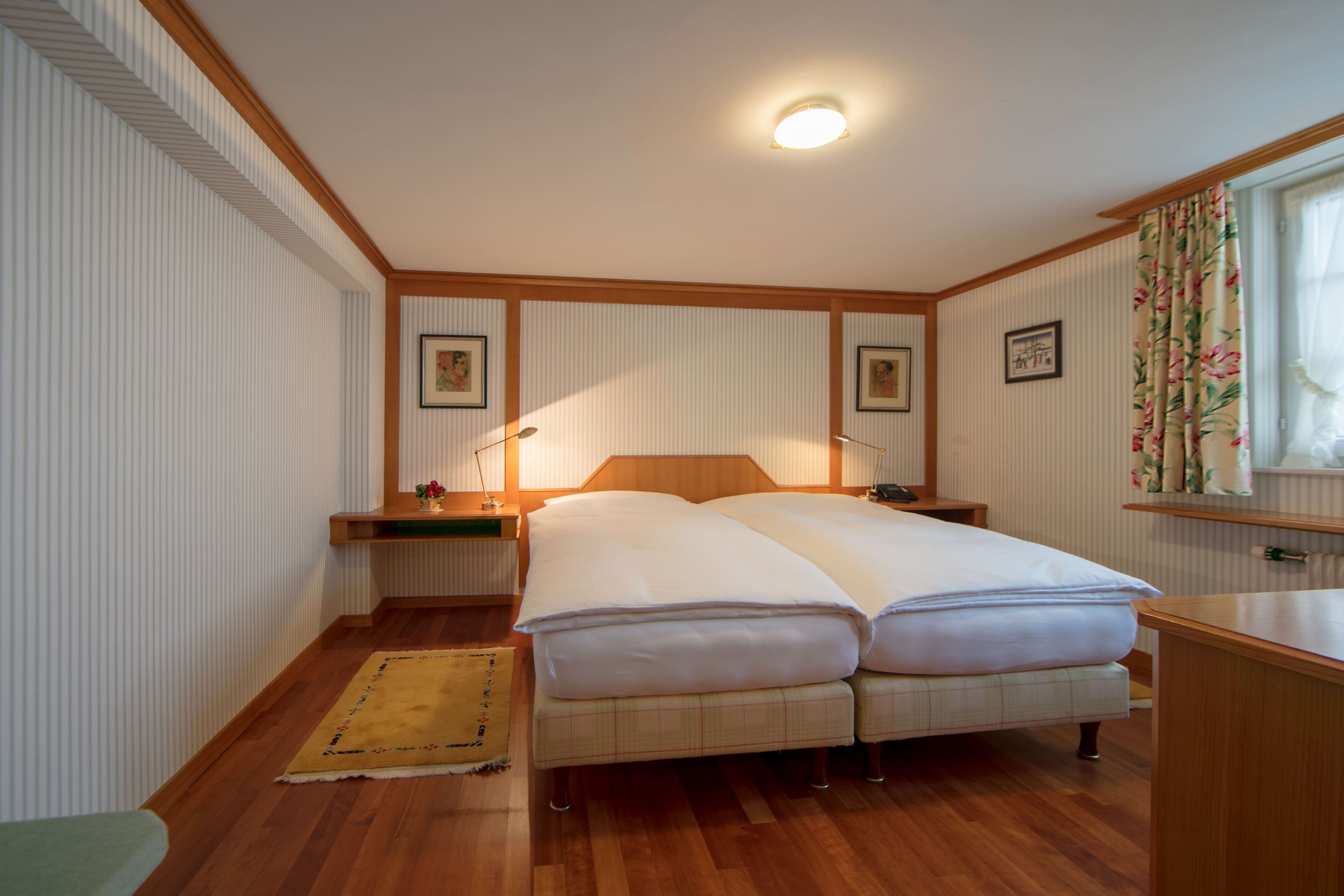 Una cama doble con cabecera de madera, una mesita de noche, un pequeño tapete y una ventana con cortinas.