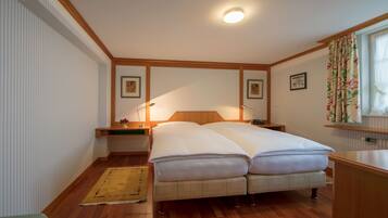Una cama doble con cabecera de madera, una mesita de noche, un pequeño tapete y una ventana con cortinas.