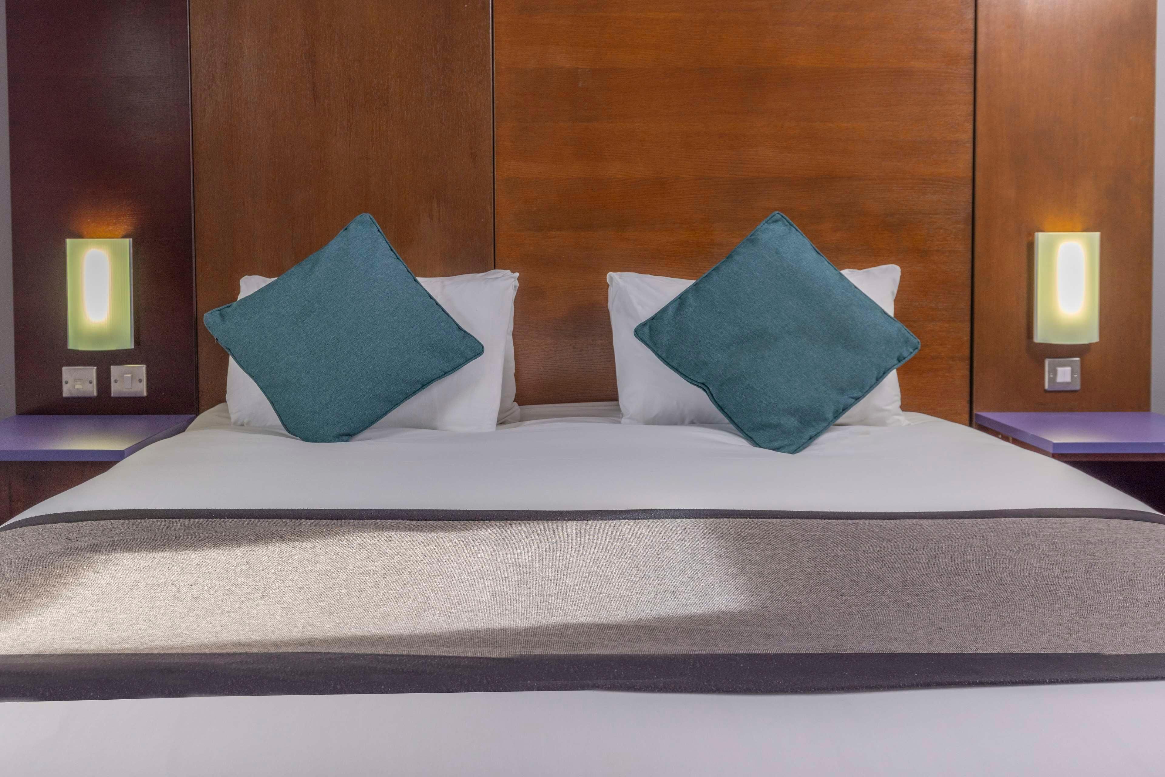 Habitación de hotel con una cama grande, dos almohadas color turquesa, una colcha blanca y cabecera de madera.