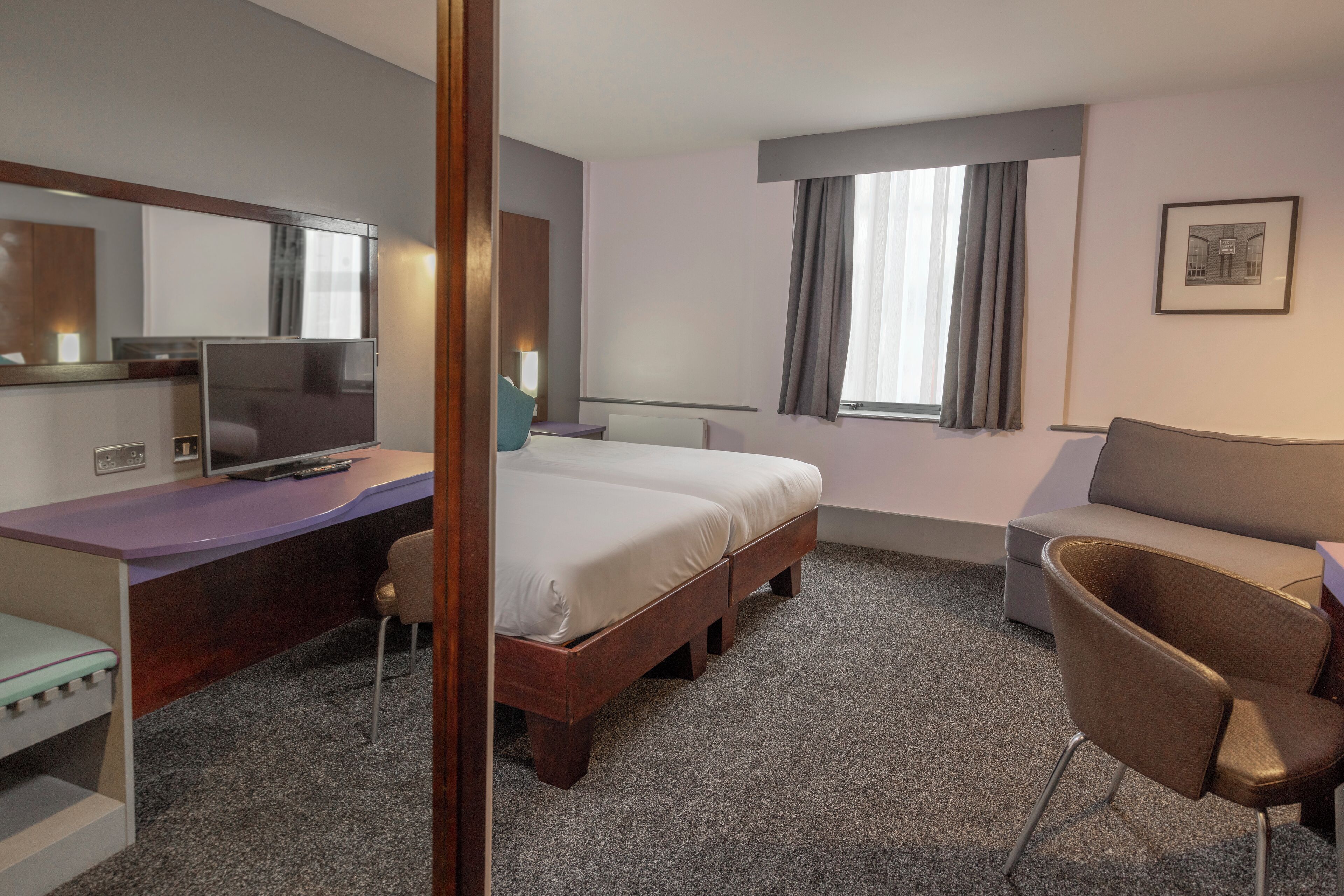 A hotel room with a bed, a desk with a TV, a chair, a sofa, and a window with curtains.