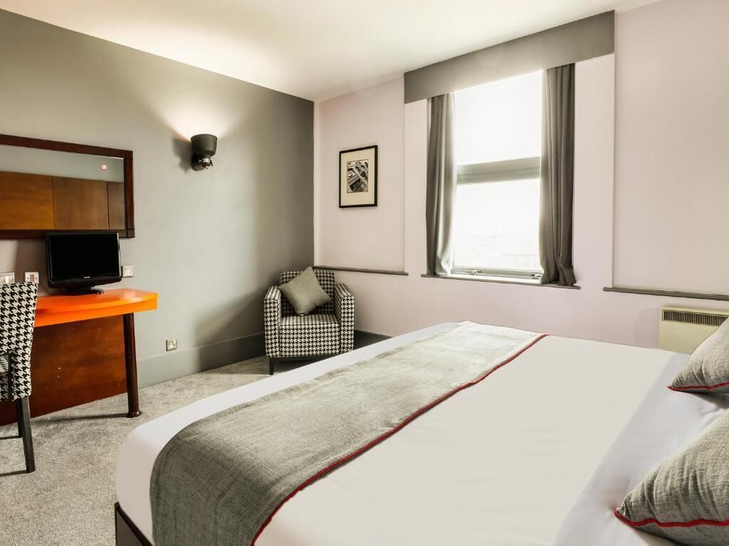 A hotel room with a bed, a desk with a television, a chair, a window with curtains, and a wall-mounted light fixture.